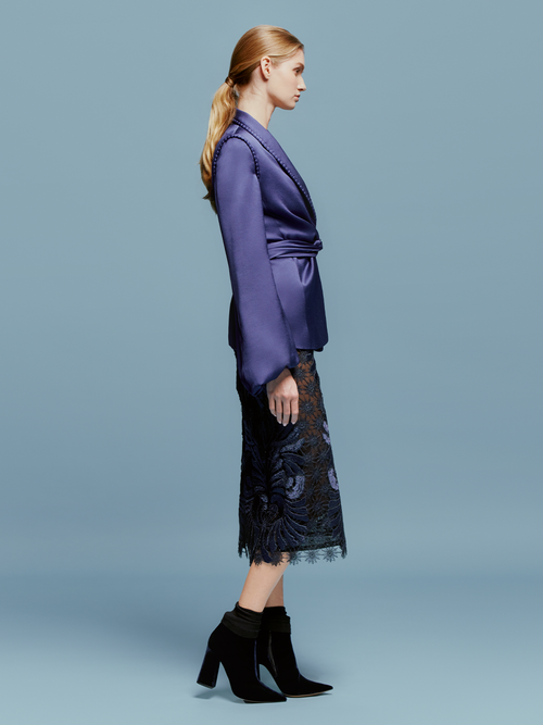 A person stands against a blue background wearing a dark purple jacket over the Kin Skirt Navy, with black heeled boots, ready for the product availability by Ship date August 2024.