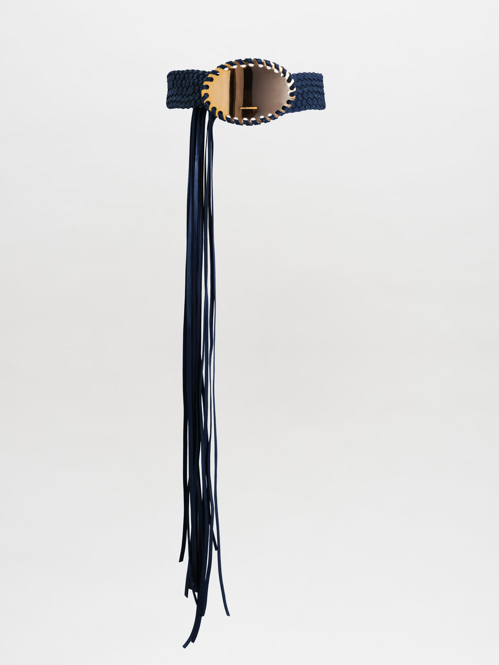 The Kai Belt is a black braided belt with a large oval gold buckle and long black fringe hanging down one side, shown on a plain light grey background.