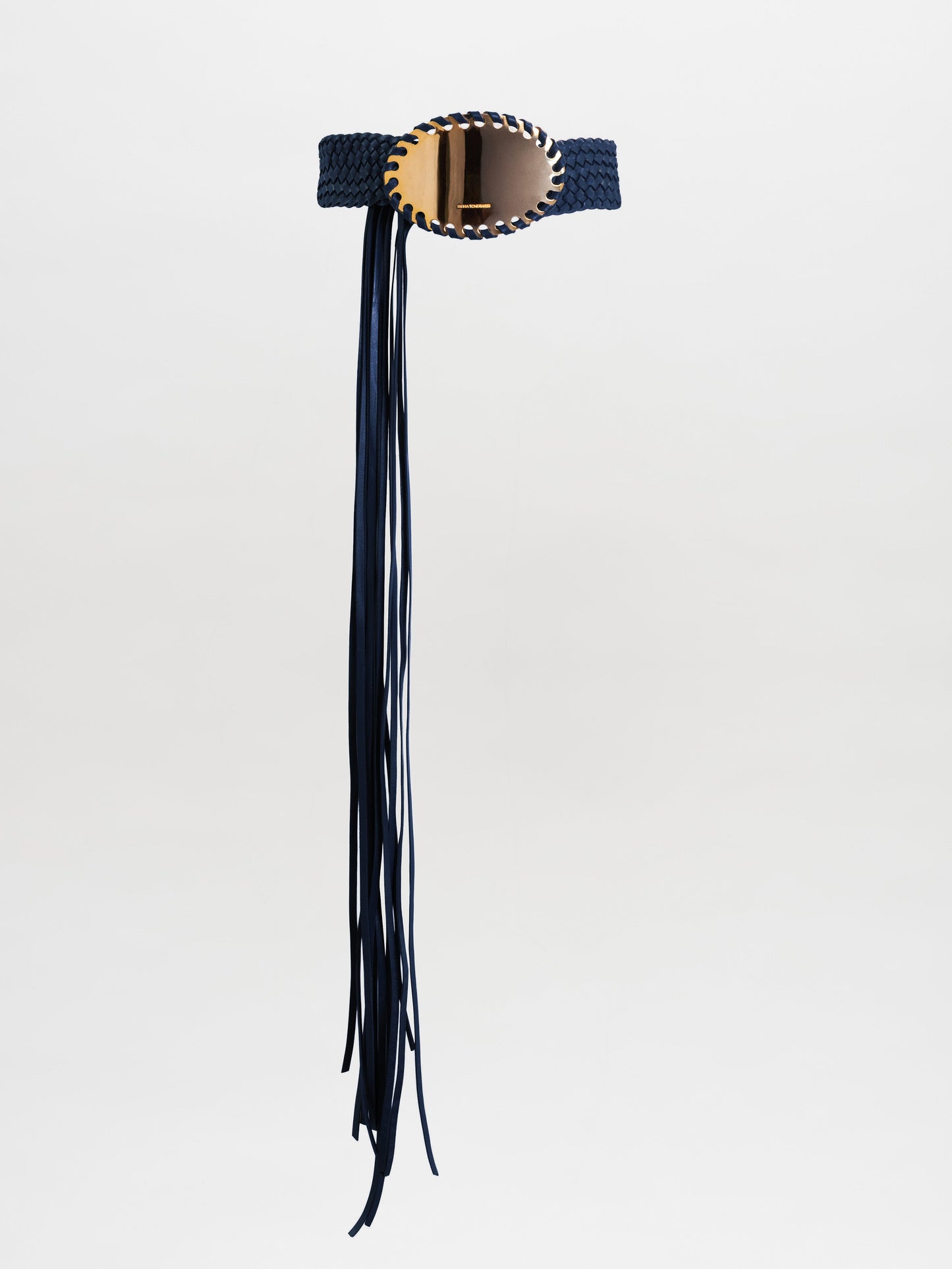 The Kai Belt is a black braided belt with a large oval gold buckle and long black fringe hanging down one side, shown on a plain light grey background.