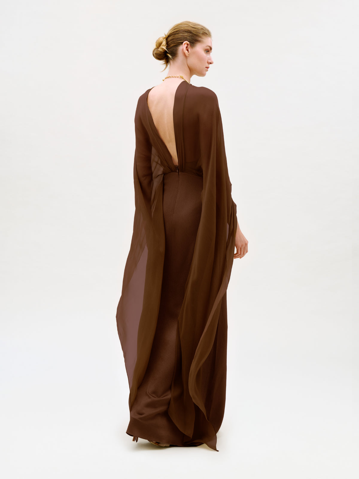 A woman with blond hair in a low bun models the Kalina Dress—a flowing, backless brown maxi featuring sheer sleeves and a graceful column silhouette—standing against a plain white background, facing away from the camera.