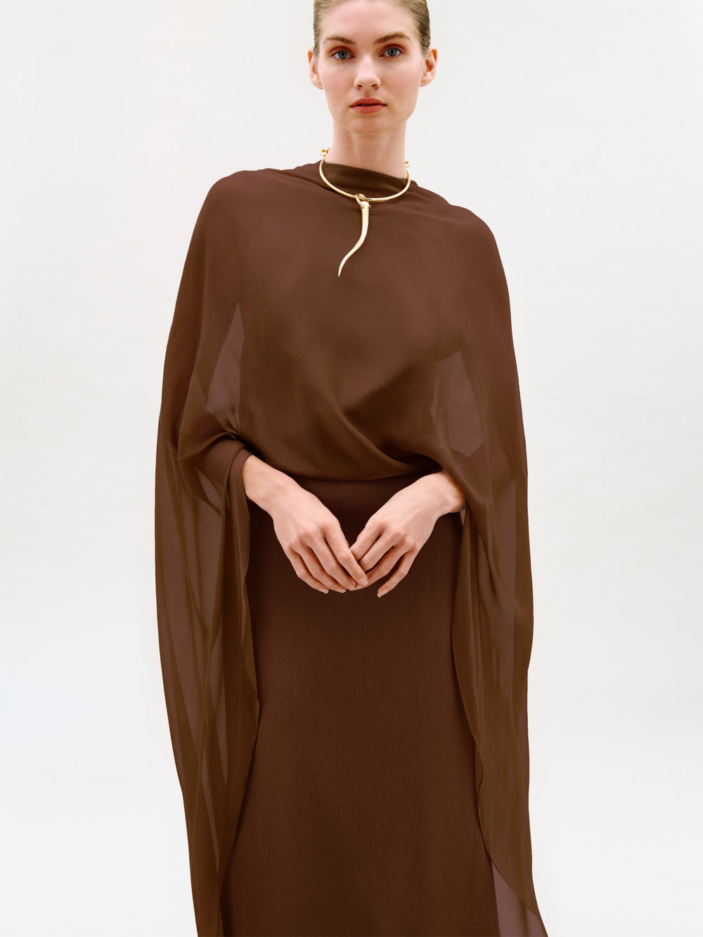 A person wearing the Kalina Dress—a sheer, brown, flowing maxi dress with long sleeves—styled with a gold necklace stands against a plain, light background. The column silhouette and luxurious Italian fabric of the Kalina Dress enhance the elegant look.