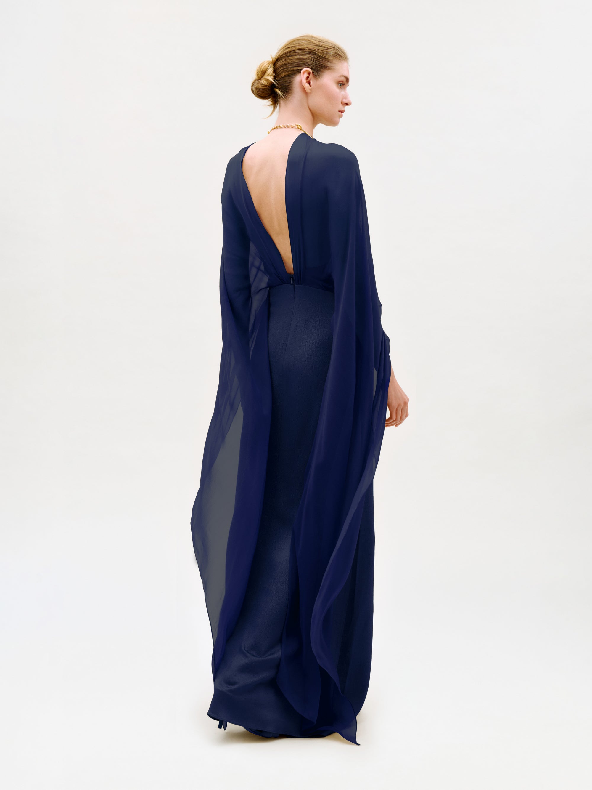 A person stands facing away, wearing the Kalina Dress—a floor-length, deep blue maxi gown crafted from luxurious Italian fabric. The dress features a sheer, flowing design with a low-cut back and column silhouette, while their hair is styled in a neat bun against a plain, light background.