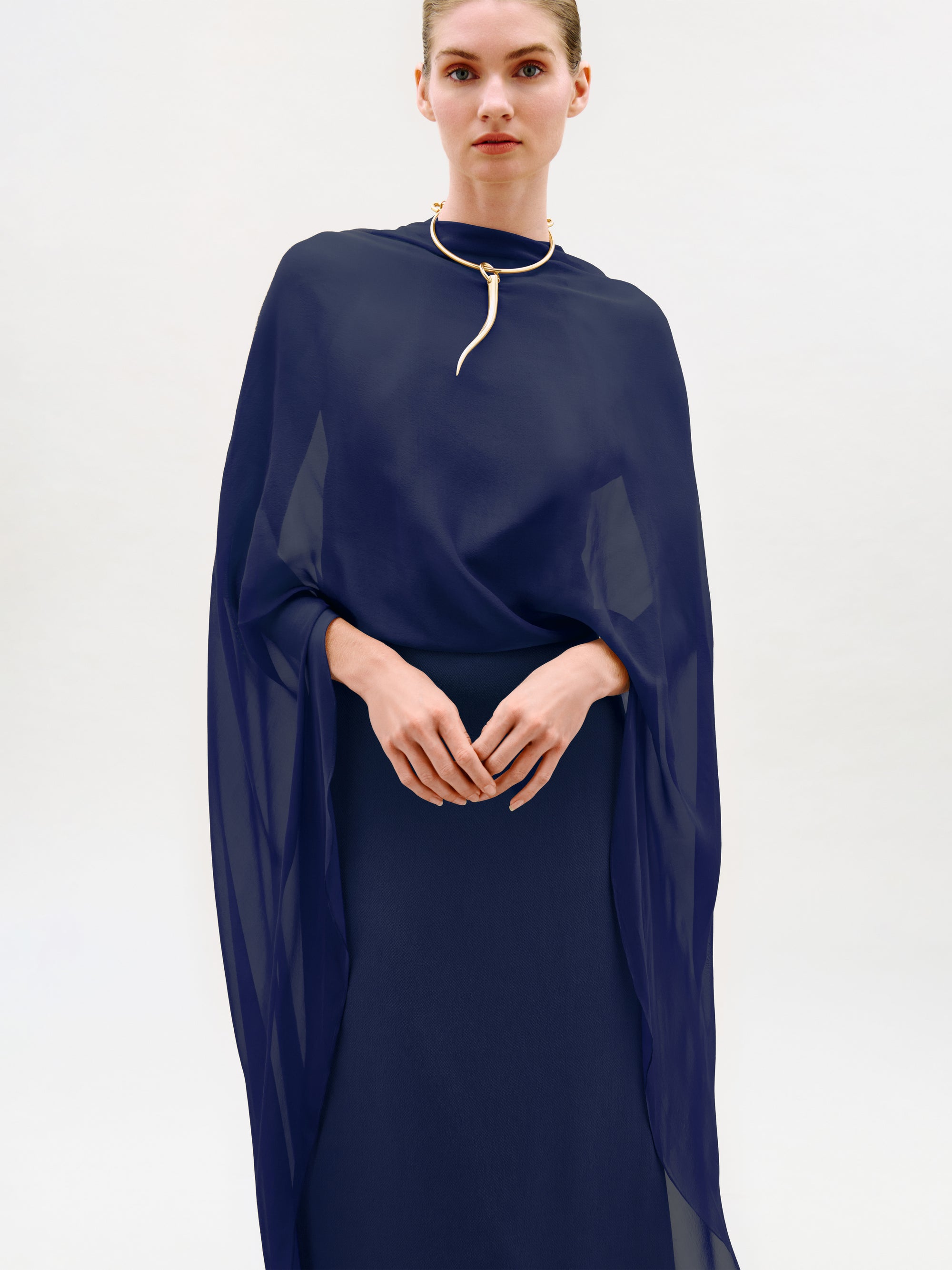 A person wearing the Kalina Dress—a sheer navy blue cape-style maxi crafted from luxurious Italian fabric—paired with a gold choker necklace, stands against a plain light background, looking at the camera with hands gently clasped.