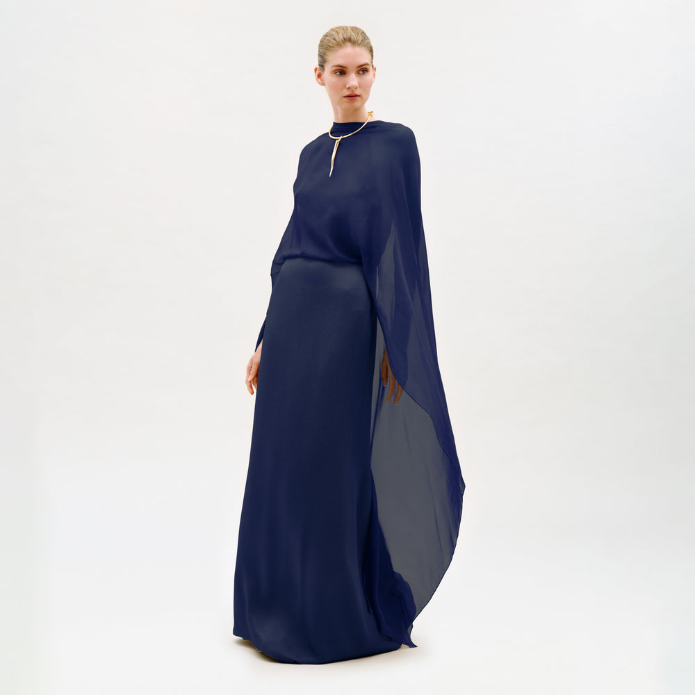 A woman stands in a studio wearing the Kalina Dress—a long, flowing navy blue maxi gown crafted from luxurious Italian fabric, with elegant cape-like sleeves and a gold necklace. The dress’s column silhouette is beautifully highlighted against the plain white background.