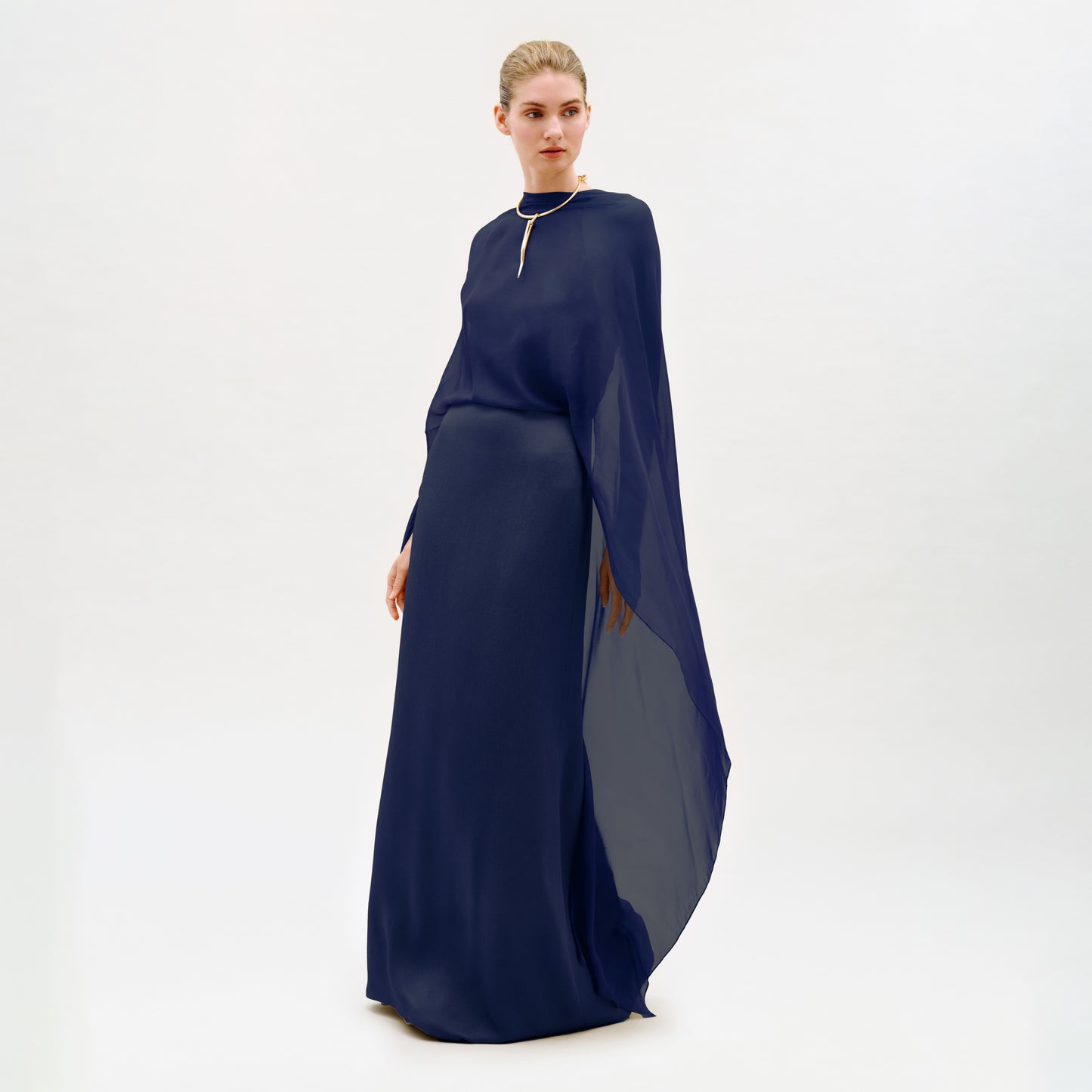A woman stands in a studio wearing the Kalina Dress—a long, flowing navy blue maxi gown crafted from luxurious Italian fabric, with elegant cape-like sleeves and a gold necklace. The dress’s column silhouette is beautifully highlighted against the plain white background.