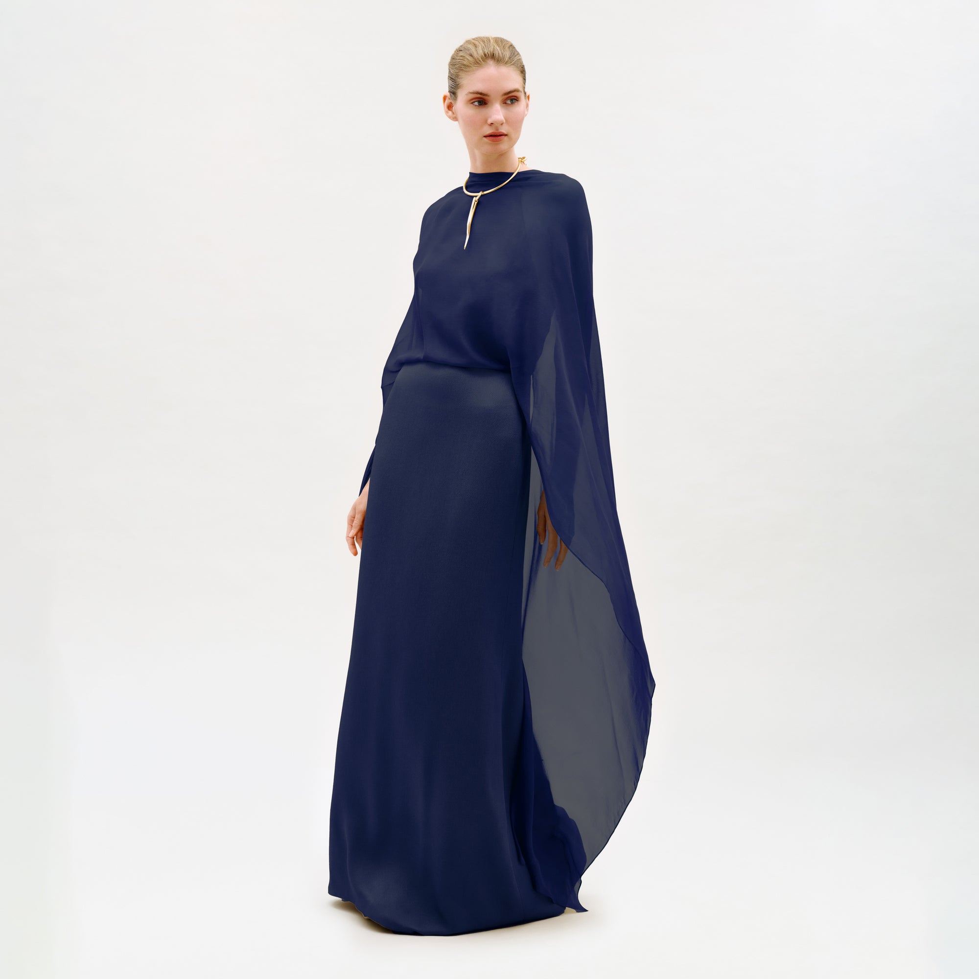 A woman stands in a studio wearing the Kalina Dress—a long, flowing navy blue maxi gown crafted from luxurious Italian fabric, with elegant cape-like sleeves and a gold necklace. The dress’s column silhouette is beautifully highlighted against the plain white background.