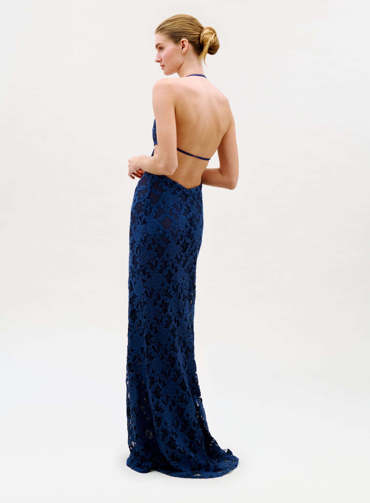 A woman in profile models the Karen Dress—a long, form-fitting navy lace gown with a halter neck and open back. Her hair is styled in a neat bun, set against a simple light background.