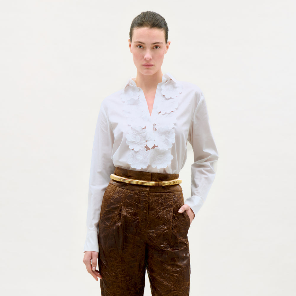 Woman wearing a white blouse with floral patterns and brown pants on a plain background