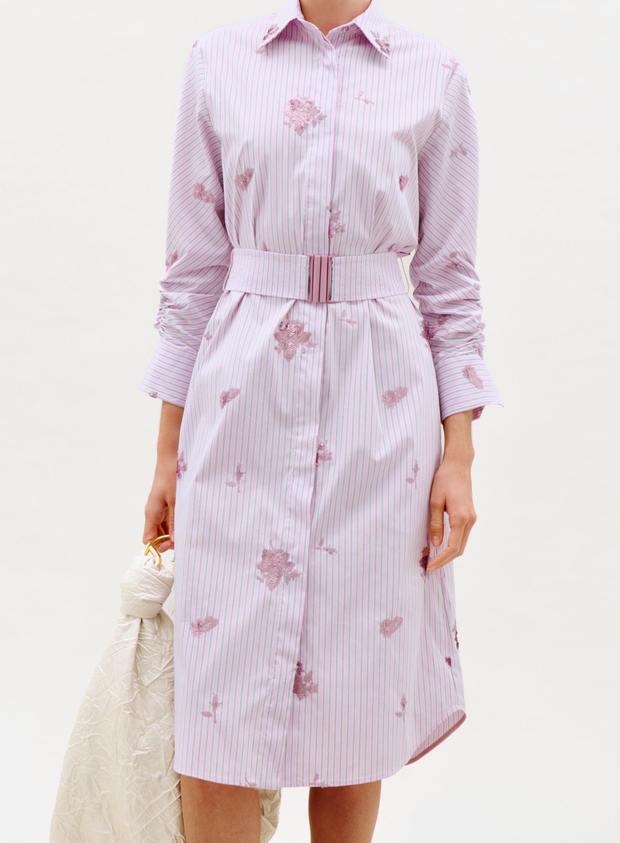 A person wears the Kendi Dress—a belted, knee-length, button-up dress with purple stripes, floral prints, and long sleeves—while holding a white textured bag against a plain white background.