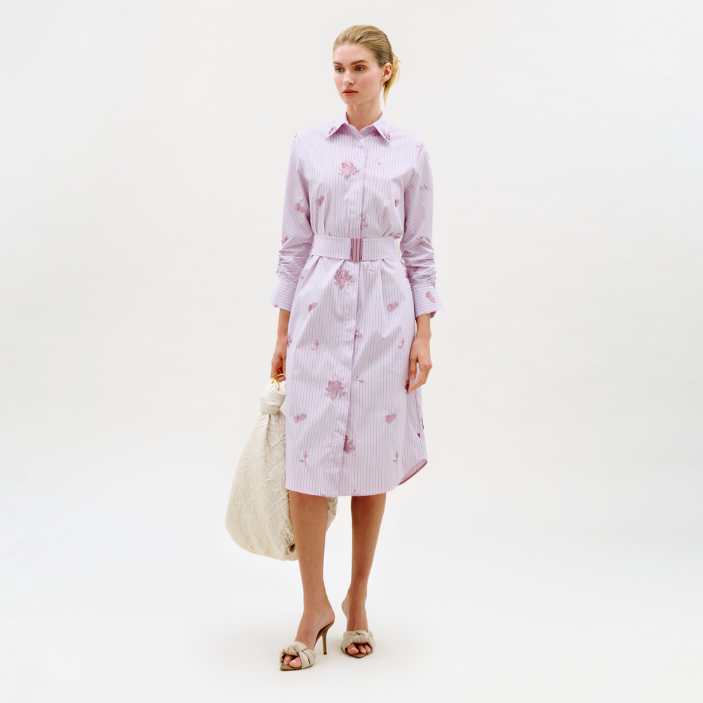 A woman wears the Kendi Dress—a long-sleeve, belted shirt dress with light pink and white stripes and floral patterns—paired with a quilted white bag and heeled sandals against a plain background.
