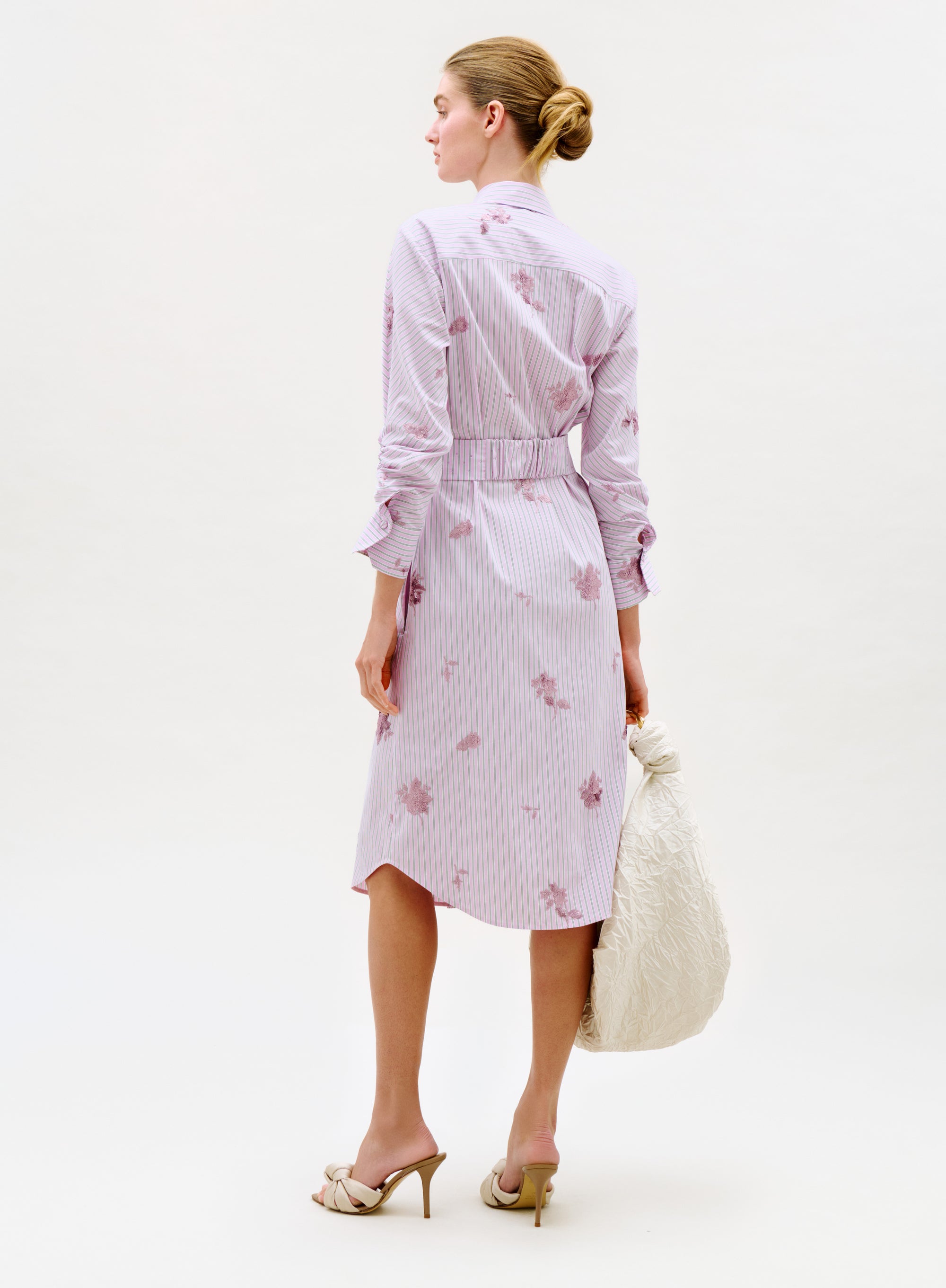 A woman stands with her back turned, wearing the Kendi Dress—a light pink striped shirt dress with floral prints. She carries a large white textured bag and wears beige high-heeled sandals. Her hair is styled in a neat bun against a plain light background.
