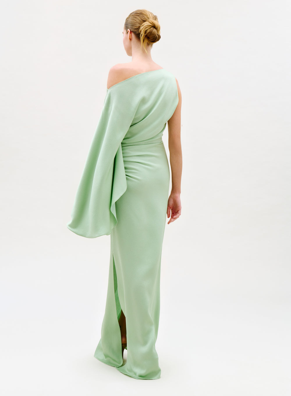 A woman faces away in the Kesia Dress, a pale green one-shoulder gown with a long draped sleeve, her hair styled in a neat bun against a plain white background.