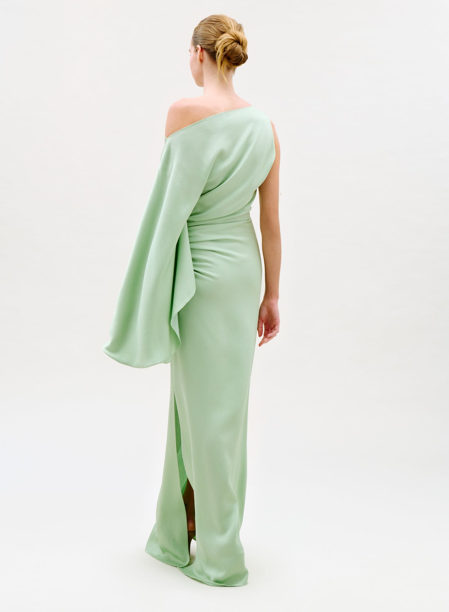 A woman faces away in the Kesia Dress, a pale green one-shoulder gown with a long draped sleeve, her hair styled in a neat bun against a plain white background.