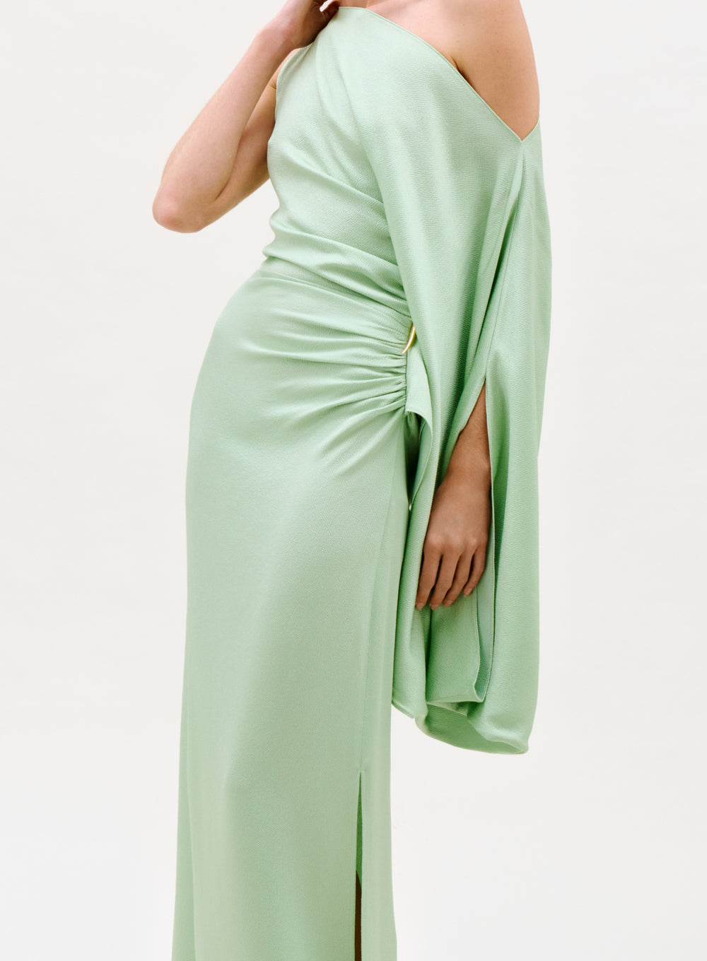 A person models the Kesia Dress—a light green, one-shoulder, floor-length gown with draped fabric and a gathered waist—against a plain white background.