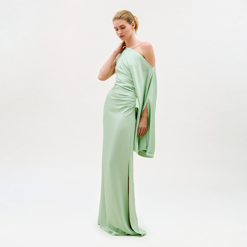 A woman wears the Kesia Dress, a light green off-the-shoulder floor-length gown, against a plain light background, looking down with one hand on her neck and the other at her side.