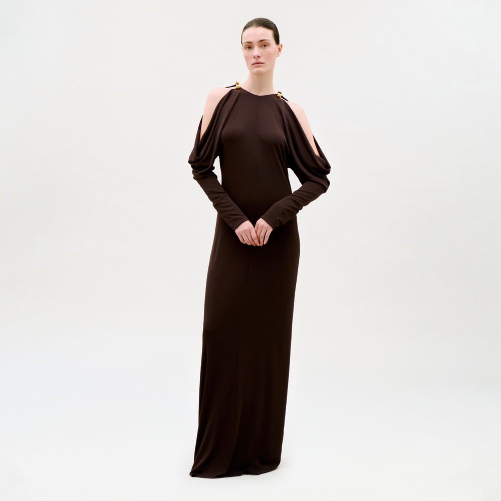 A woman stands against a plain light background, wearing the Kilian Dress—a long, dark brown dress featuring cold shoulder cutouts and gold-accented shoulders. Her hair is pulled back and her hands are clasped in front of her.