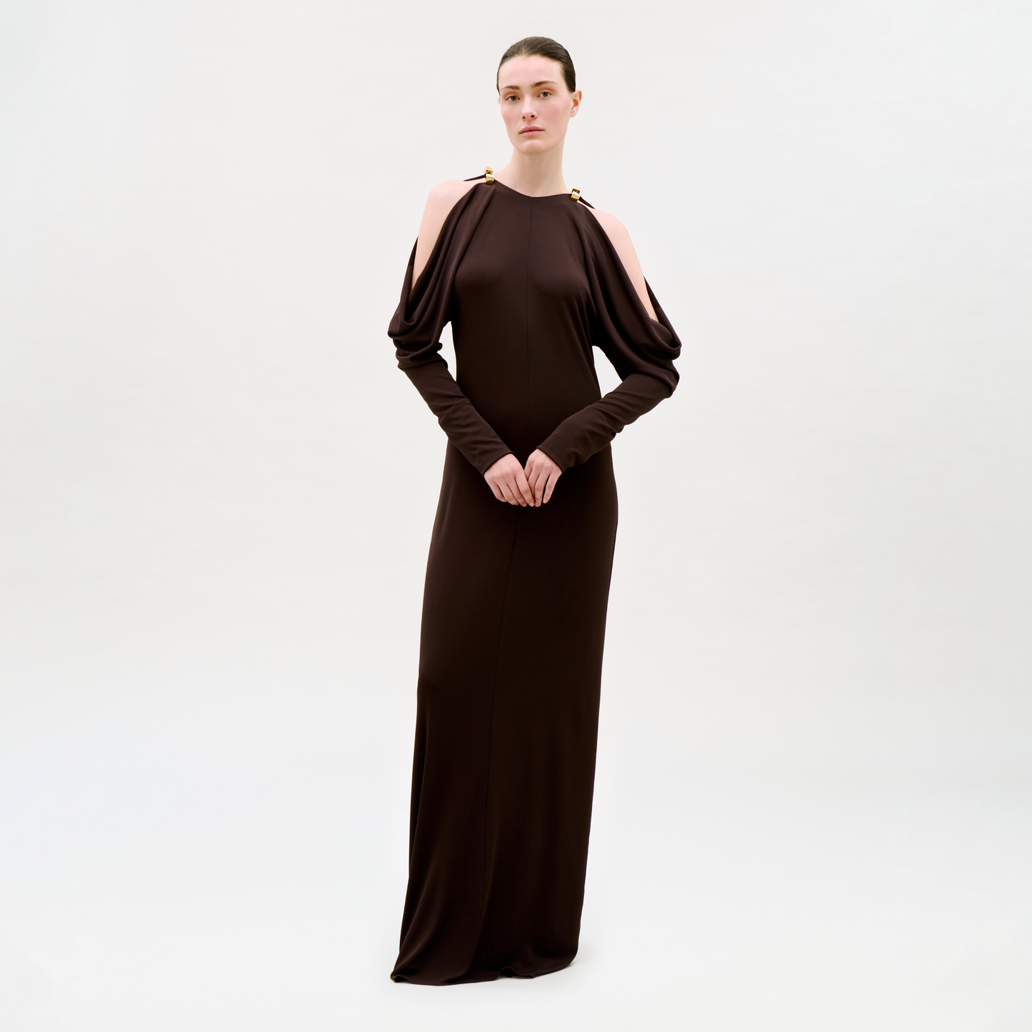 A woman stands against a plain light background, wearing the Kilian Dress—a long, dark brown dress featuring cold shoulder cutouts and gold-accented shoulders. Her hair is pulled back and her hands are clasped in front of her.