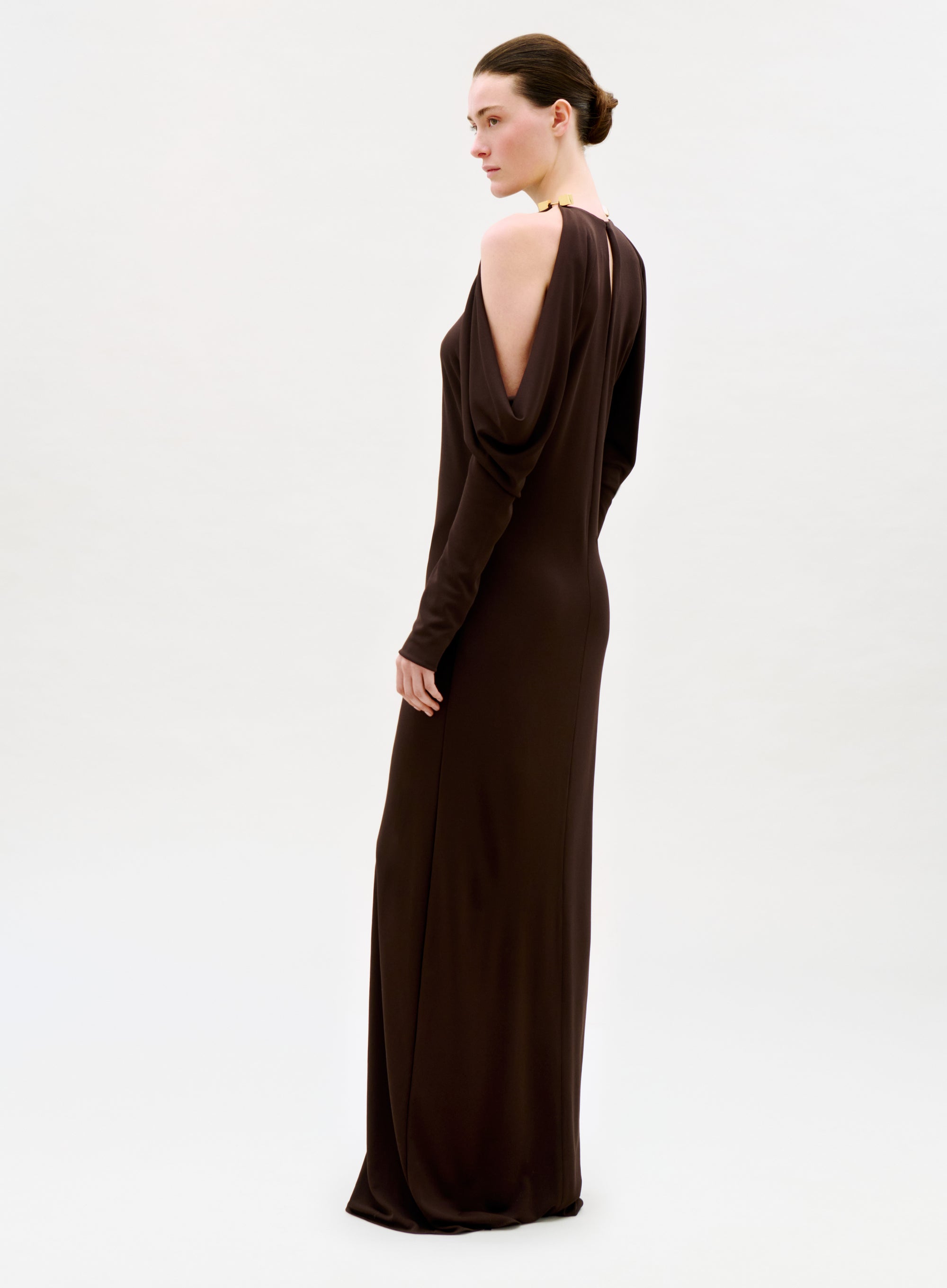 A woman in profile wears the Kilian Dress—a long, dark brown gown with draped and open shoulder details. Her hair is neatly pulled back in a bun as she poses against a simple, light background.