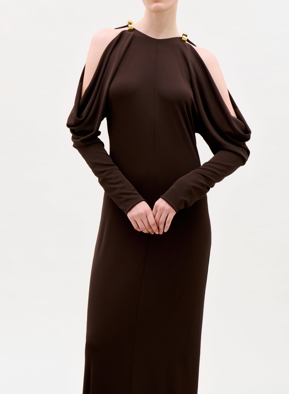 A person wearing the Kilian Dress, a long dark brown gown with draped cold-shoulder sleeves and gold clasp details at the shoulders, stands against a plain light background.