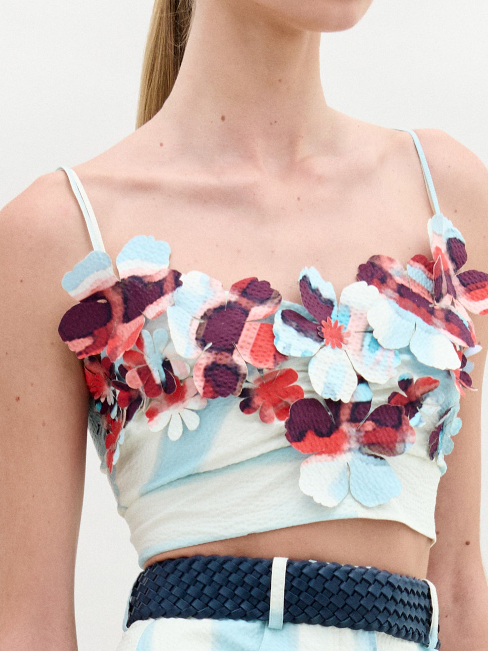 A woman stands against a plain background wearing the Kim Top— a sleeveless crop top with striking 3D floral detailing in red and blue—paired with a light blue patterned skirt.