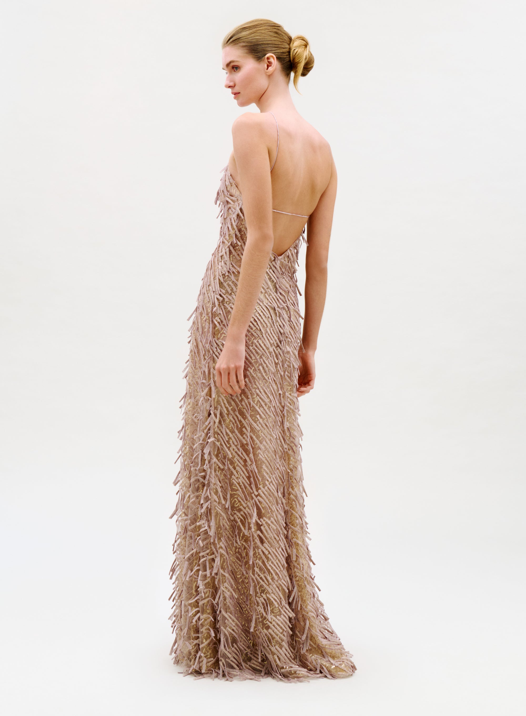 A woman stands in profile against a plain light background, her hair in a neat low bun, wearing the Kora Dress—a backless, floor-length nude gown with textured feather-like details.
