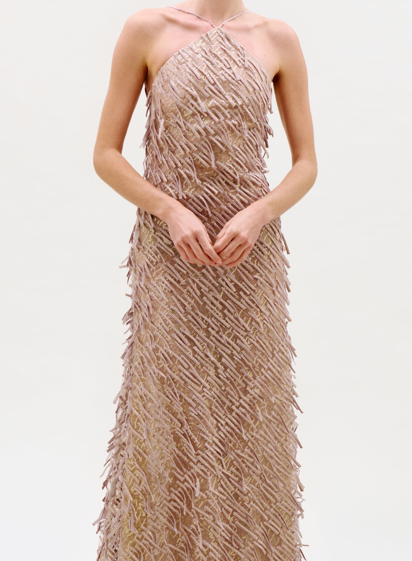 A woman wearing the Kora Dress, a sleeveless, textured champagne gown with a halter neckline, stands against a plain light background with her hands gently clasped at her waist.