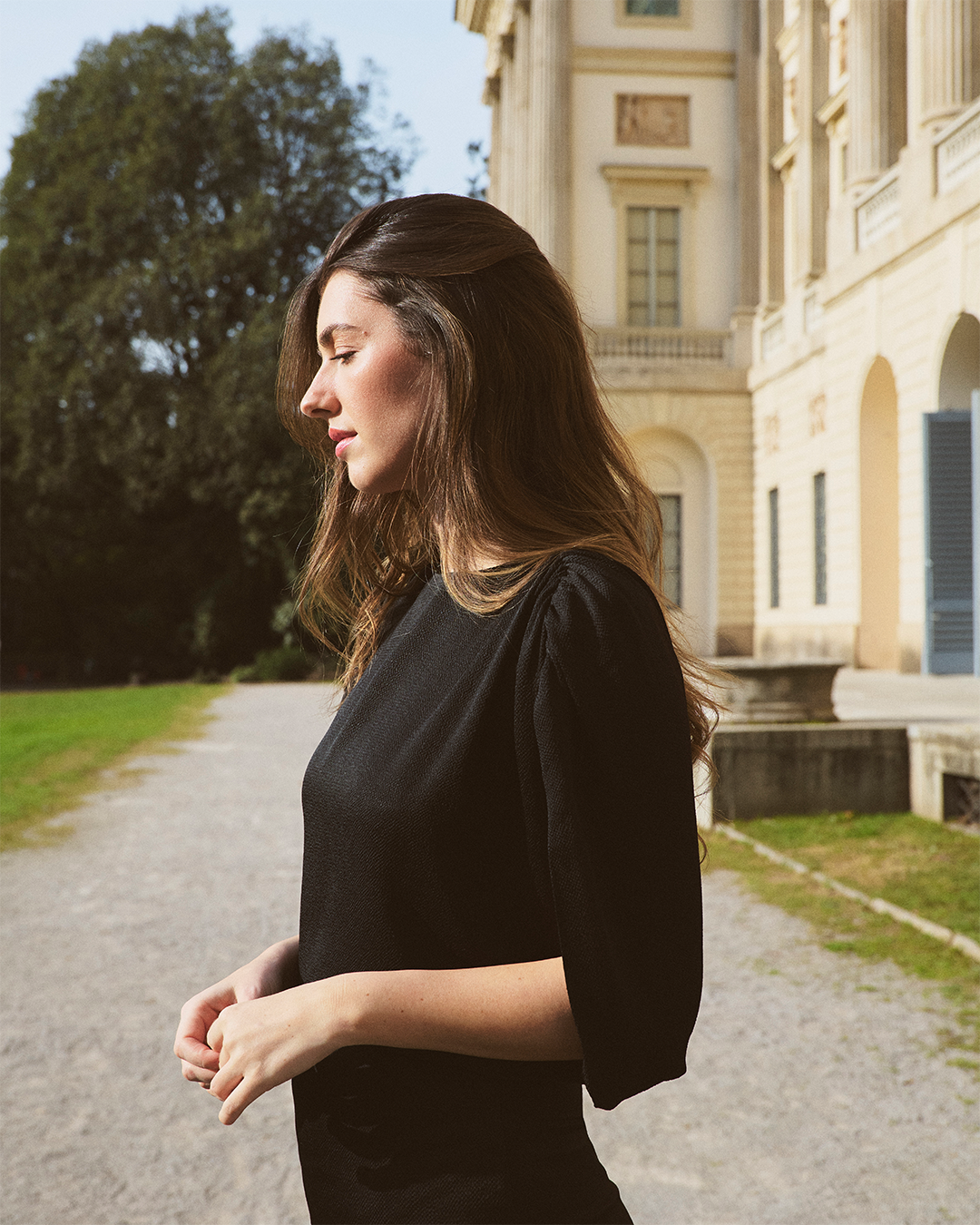 A woman with long hair in a black dress stands outdoors near a historic building with arches and green trees.