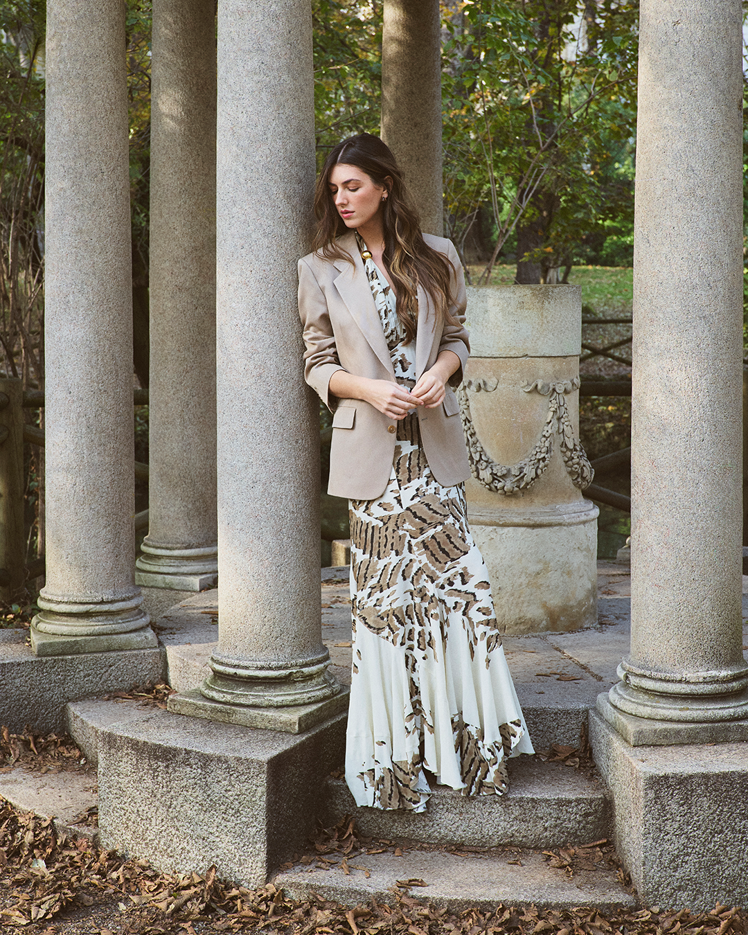 A woman in a patterned dress and beige blazer stands among stone columns in a leafy outdoor setting.