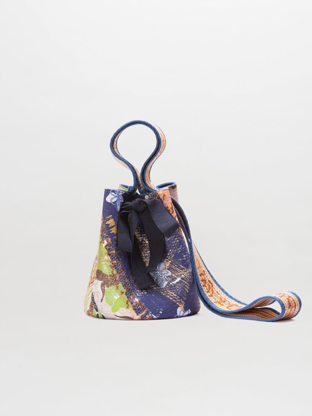 The Mochila Laaput Small is a hand-woven Wayuu bucket bag featuring a multicolored abstract pattern, black bow detail, and shoulder strap, displayed on a plain white background.