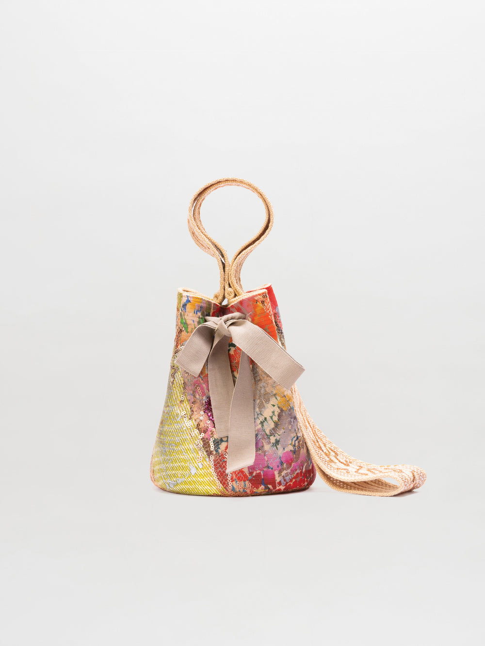 The Mochila Laaput Small is a handmade patchwork bucket bag with a large beige bow, braided handle, and long woven strap, displayed against a plain light background.