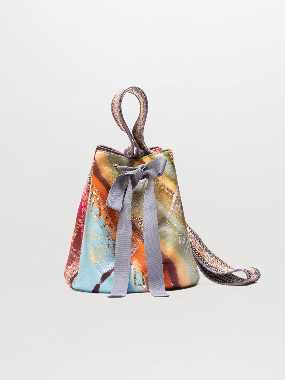 The Mochila Laaput Big is a multicolored Wayuu mochila bucket bag with abstract patterns, a gray ribbon at the front, and a hand-woven patterned shoulder strap, shown against a plain background.