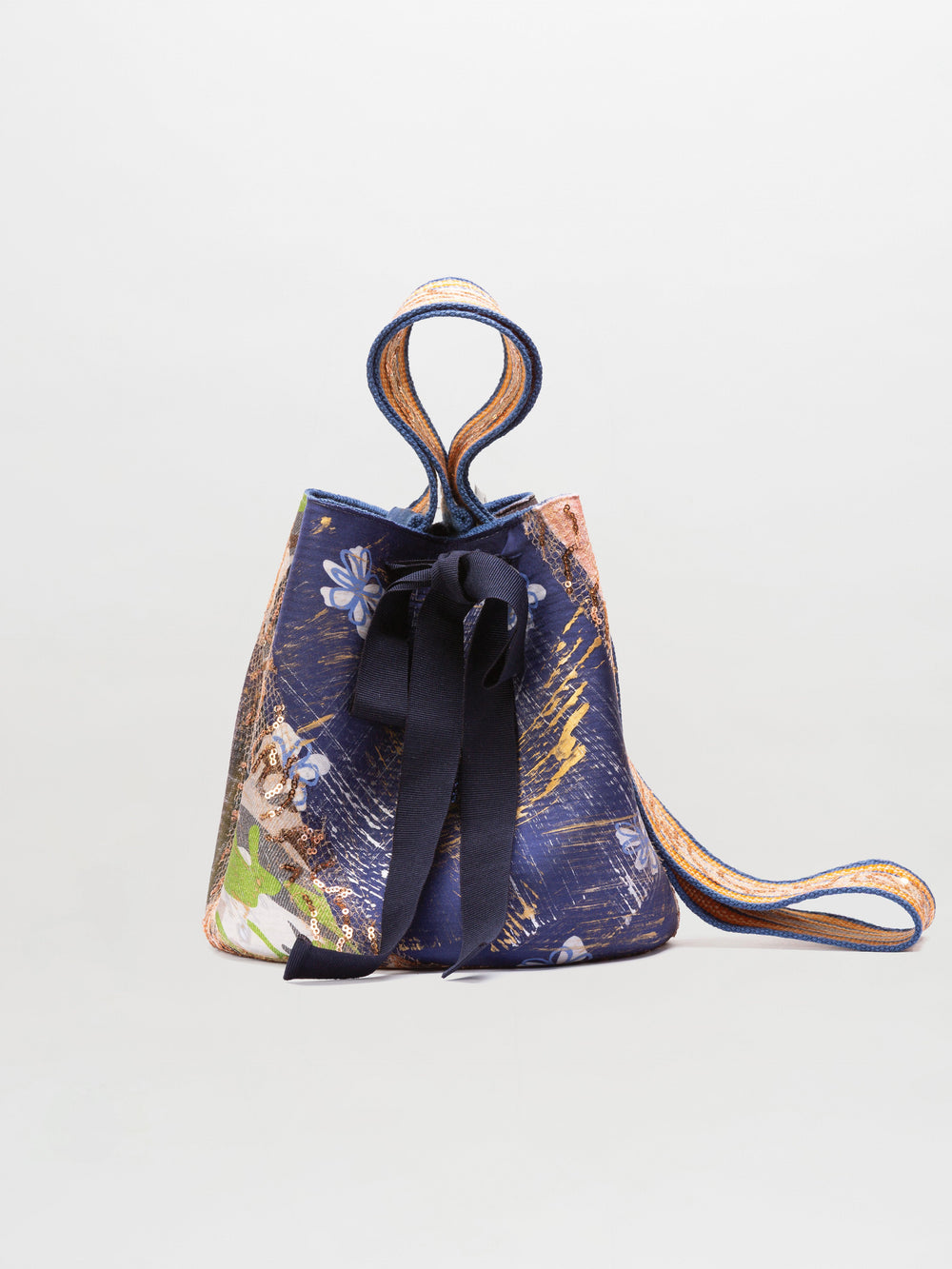The Mochila Laaput Big is a hand-woven bucket handbag with a blue floral and abstract pattern, black front ribbon, and long striped shoulder strap, shown on a plain background.