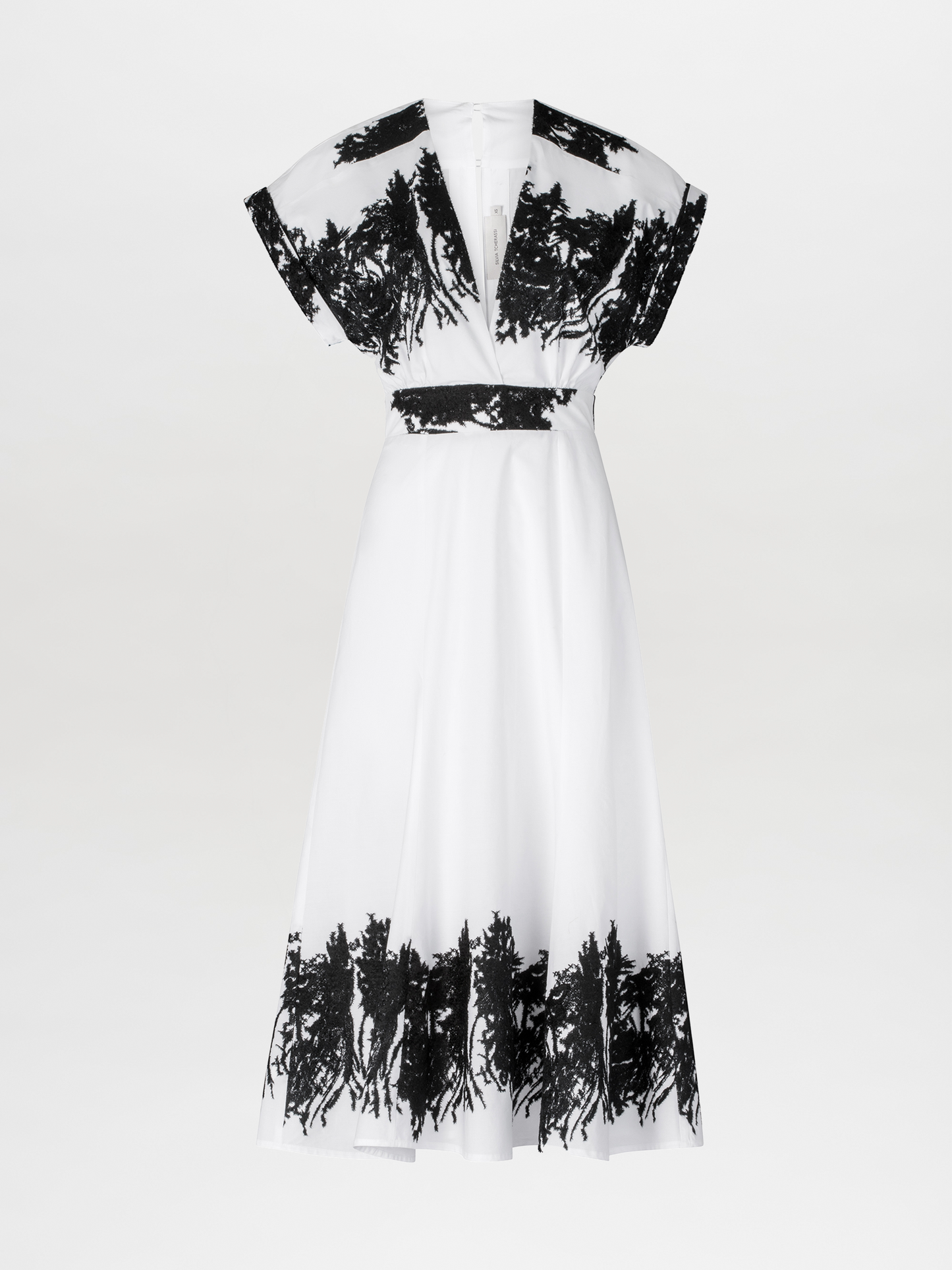 The individual is wearing the Laila Dress, a white midi dress adorned with black patterns and embroidered floral details, set against a light blue backdrop. The dress boasts a chic V-neckline and is stylishly paired with white ankle boots and a belt.