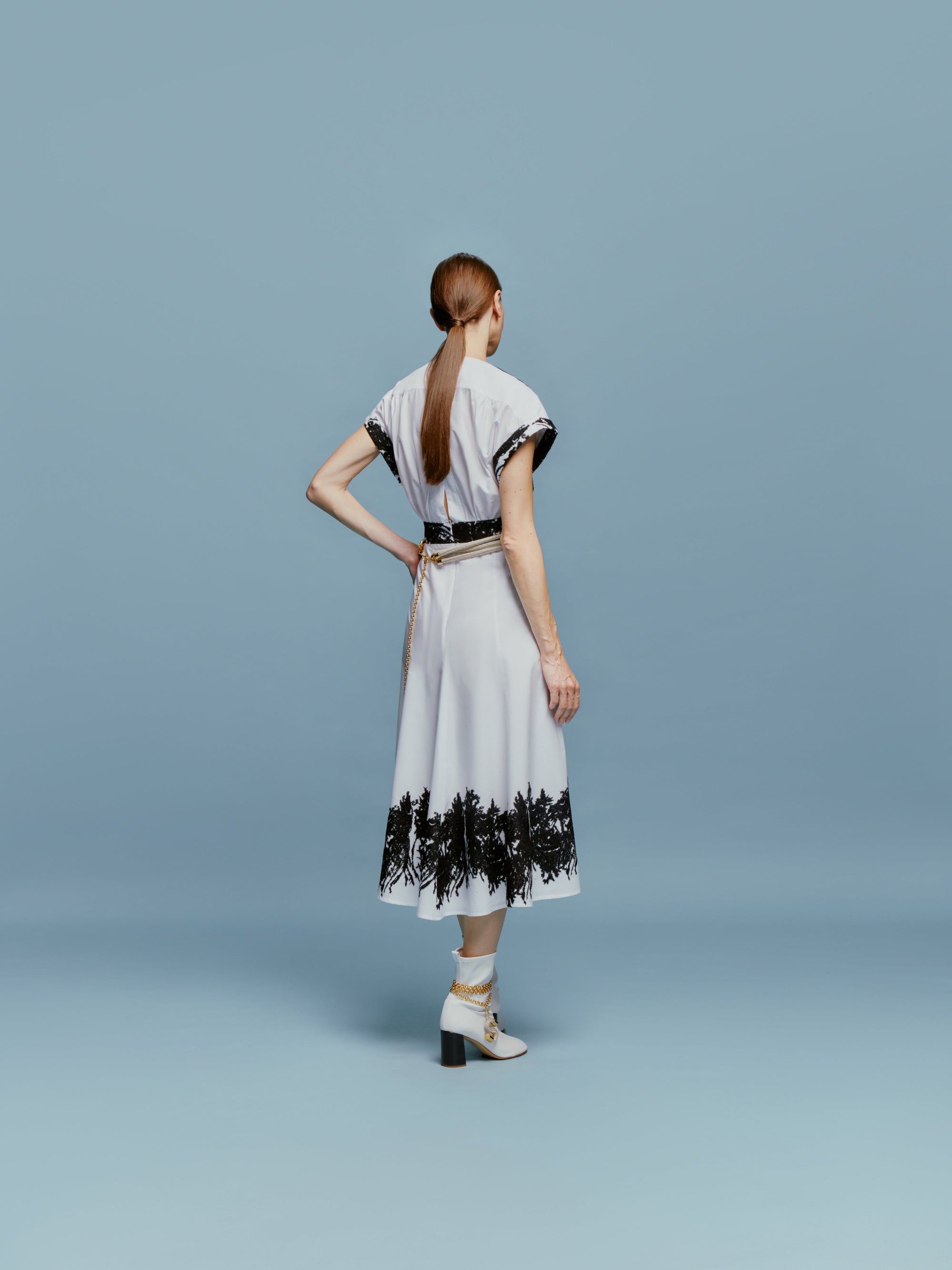 The Laila Dress features a white canvas with contrasting patterns, embroidered black floral details on the sleeves, waist, and hem, and is complemented by a V-neck and short sleeves against a plain backdrop.