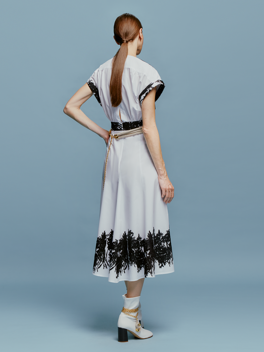 On September 2nd, a woman stands against a light blue background wearing the Laila Dress White Black Velvet Embroidery, paired with white ankle boots and a beige belt with hanging tassels.