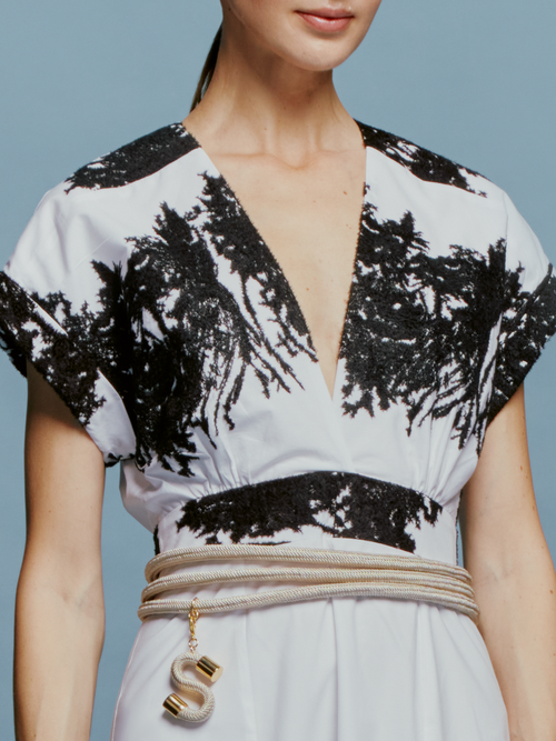 On September 2nd, a woman stands against a light blue background wearing the Laila Dress White Black Velvet Embroidery, paired with white ankle boots and a beige belt with hanging tassels.