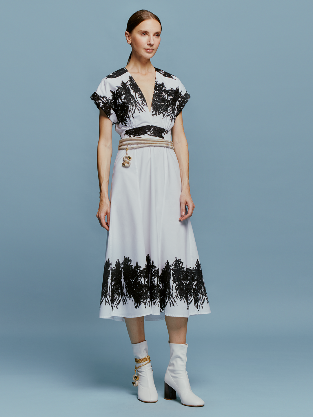 On September 2nd, a woman stands against a light blue background wearing the Laila Dress White Black Velvet Embroidery, paired with white ankle boots and a beige belt with hanging tassels.