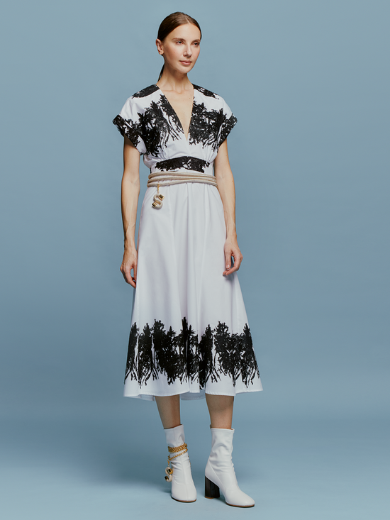 On September 2nd, a woman stands against a light blue background wearing the Laila Dress White Black Velvet Embroidery, paired with white ankle boots and a beige belt with hanging tassels.