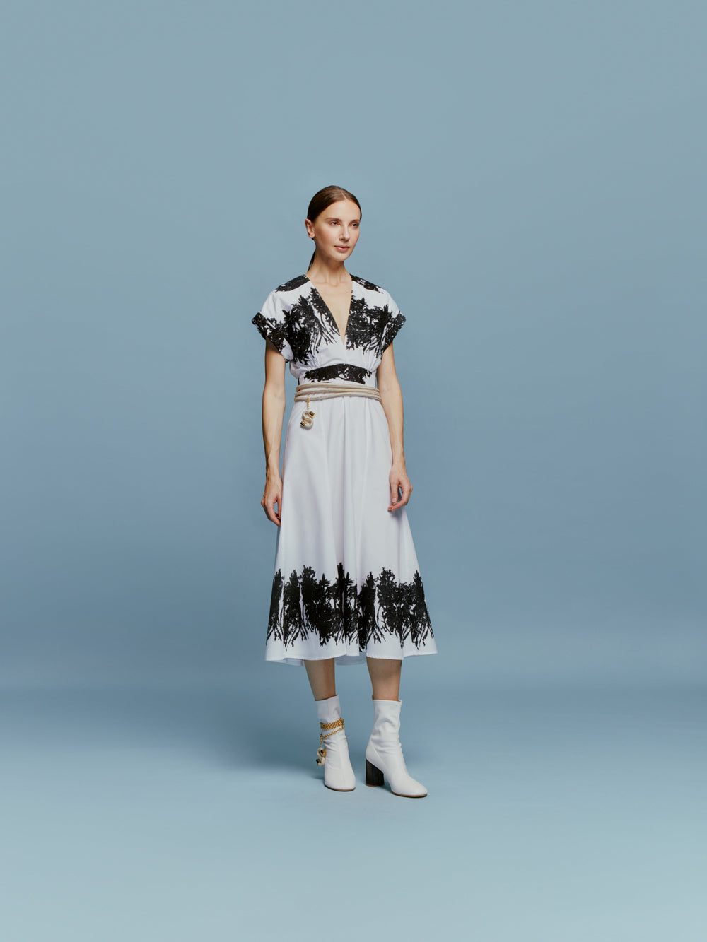 The Laila Dress features a white canvas with contrasting patterns, embroidered black floral details on the sleeves, waist, and hem, and is complemented by a V-neck and short sleeves against a plain backdrop.