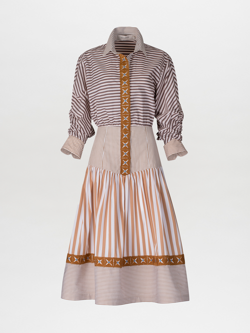 A woman stands against a blue background, wearing the Lamar Dress in Multi Brown Stripes, accentuated with a belt and boots, evoking the spirit of an August 19th summer day.