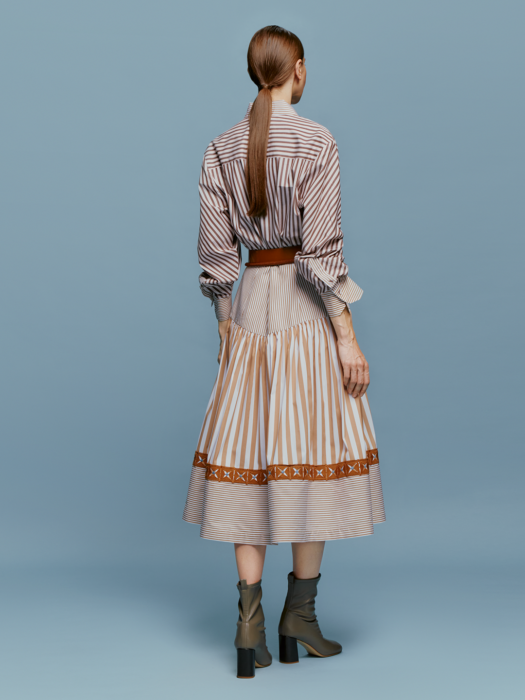 A woman stands against a blue background, wearing the Lamar Dress in Multi Brown Stripes, accentuated with a belt and boots, evoking the spirit of an August 19th summer day.