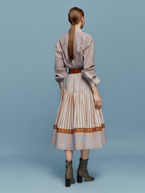 A woman stands against a blue background, wearing the Lamar Dress in Multi Brown Stripes, accentuated with a belt and boots, evoking the spirit of an August 19th summer day.