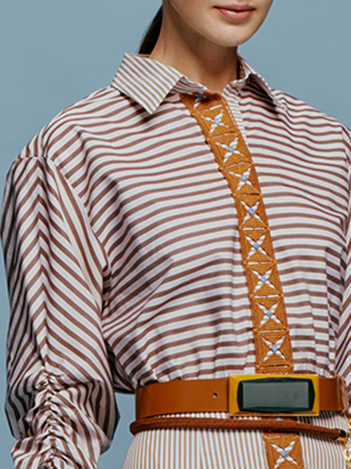 A woman stands against a blue background, wearing the Lamar Dress in Multi Brown Stripes, accentuated with a belt and boots, evoking the spirit of an August 19th summer day.