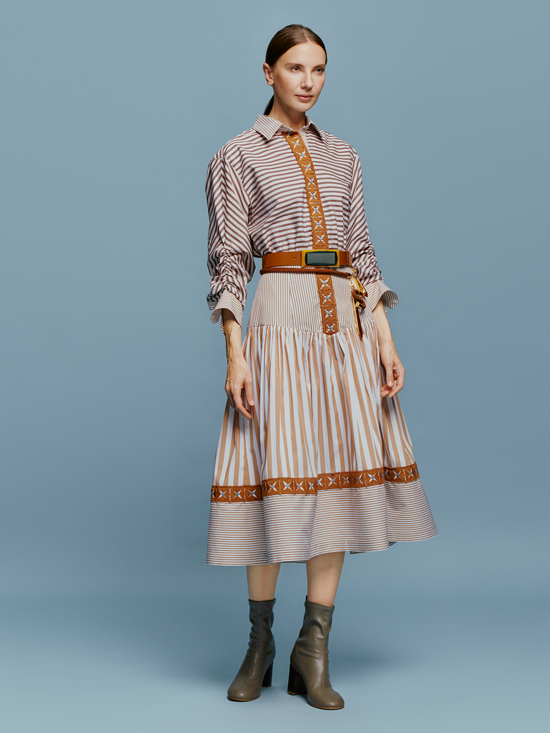 A woman stands against a blue background, wearing the Lamar Dress in Multi Brown Stripes, accentuated with a belt and boots, evoking the spirit of an August 19th summer day.