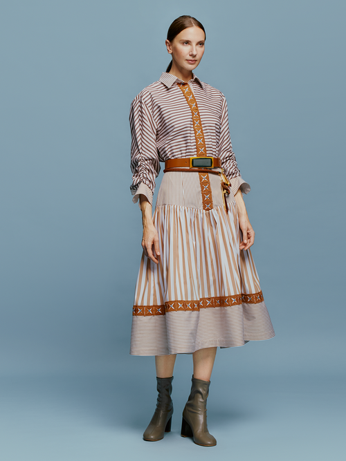 A woman stands against a blue background, wearing the Lamar Dress in Multi Brown Stripes, accentuated with a belt and boots, evoking the spirit of an August 19th summer day.