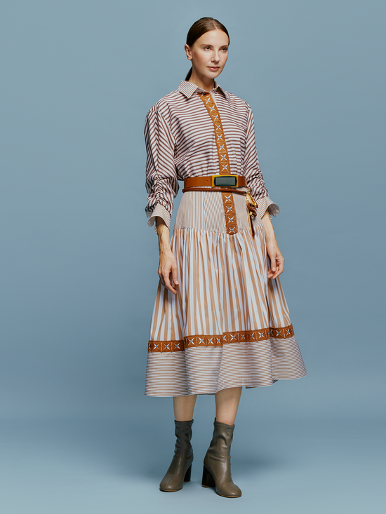 A woman stands against a blue background, wearing the Lamar Dress in Multi Brown Stripes, accentuated with a belt and boots, evoking the spirit of an August 19th summer day.