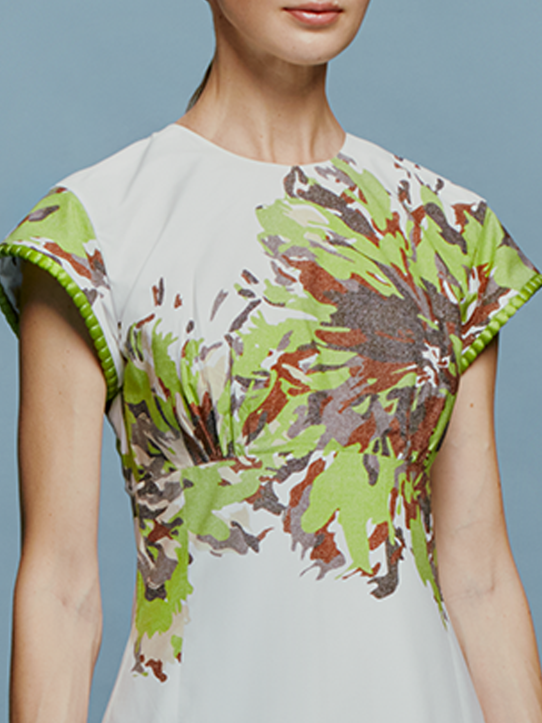 A woman in the Lara Dress Floral Camo, featuring a white base with green floral patterns, stands against a blue background. She is wearing beige high-heeled shoes and wooden bangles, perfectly capturing the essence of August 19th.