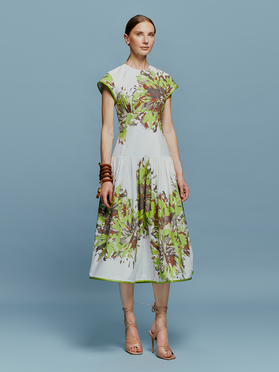 A woman in the Lara Dress Floral Camo, featuring a white base with green floral patterns, stands against a blue background. She is wearing beige high-heeled shoes and wooden bangles, perfectly capturing the essence of August 19th.