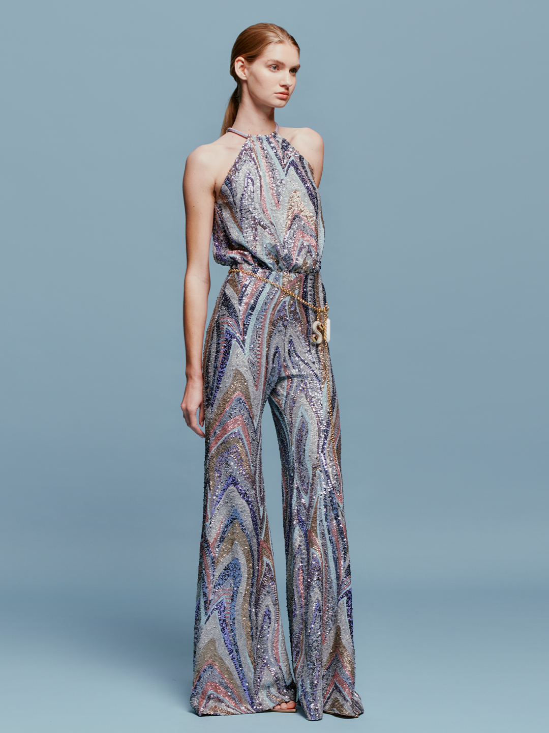 Against a plain blue background, a person stands poised in the Lauretta Jumpsuit Sequin Waves, featuring a sleeveless, high-neck design with a colorful, swirling pattern. The outfit showcases wide-leg pants and comes with a belt—perfectly capturing the style for upcoming events between August 19th and September 2nd, 2024.