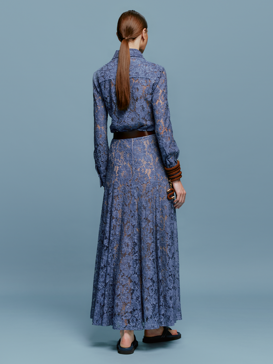 A woman stands against a blue background wearing the Lavanda Skirt Blue paired with a long-sleeved, blue lace top, accessorized with a brown belt, beaded necklace, and brown bangles. She has her hair tied back and is wearing black sandals. This stylish look will be available starting August 19th, 2024.